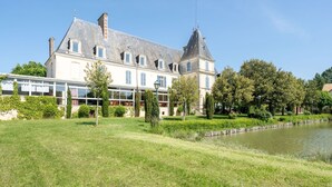 Un edificio grande e histórico con una torre destacada, un patio con un estanque y abundante vegetación bien cuidada.