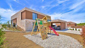 Un edificio moderno con zona de juegos que incluye un tobogán y pared de escalar, rodeado de grava y vegetación.
