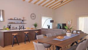 Una cocina moderna con una isla central, taburetes de bar, una mesa de comedor con sillas, un sofá y un televisor al fondo.