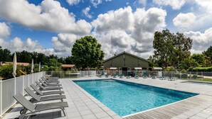 A rectangular swimming pool with a tiled deck, surrounded by lounge chairs and umbrellas, with a building and trees in the background. - Serene Chalet in Hourtin near Lake (Hourtin)