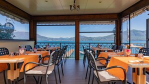 Un restaurant doté d’une terrasse offrant une vue sur un plan d’eau et des montagnes. Les tables sont disposées avec des chaises, et l’espace est délimité par une rambarde.