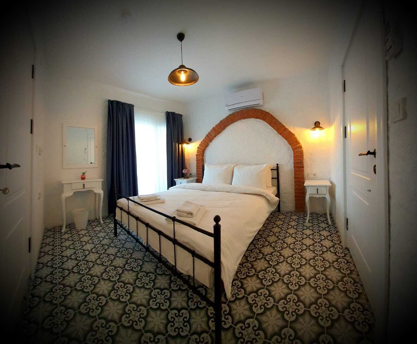Quarto com cama, mesa de cabeceira, luminária pendente, ar condicionado e piso com ladrilhos estampados.