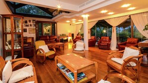 A spacious living room with wooden furniture, a glass cabinet, and large windows offering a view of the outdoors.