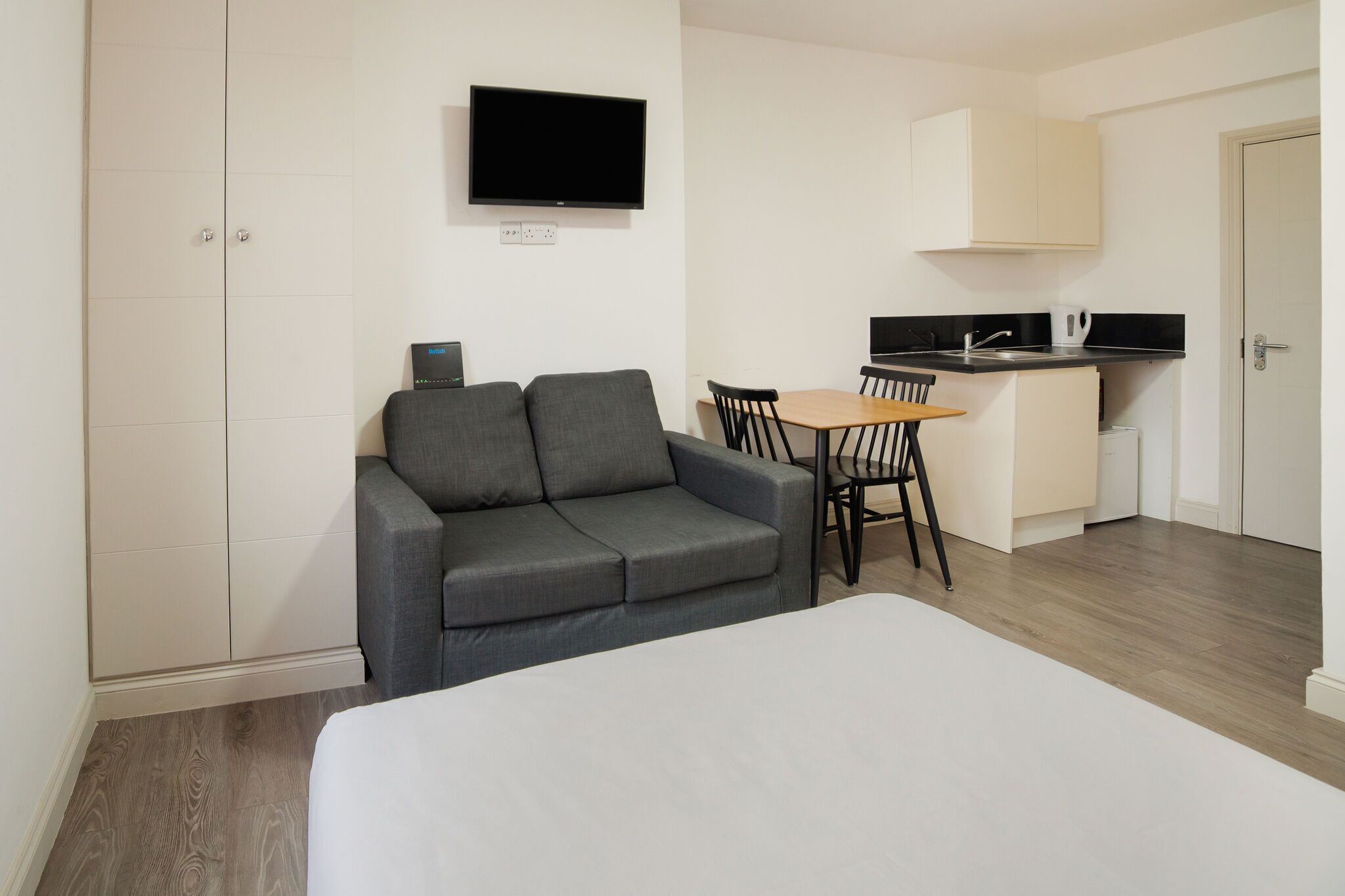 A room with a bed, a grey sofa, a small table with chairs, a TV mounted on the wall, and a kitchenette area.