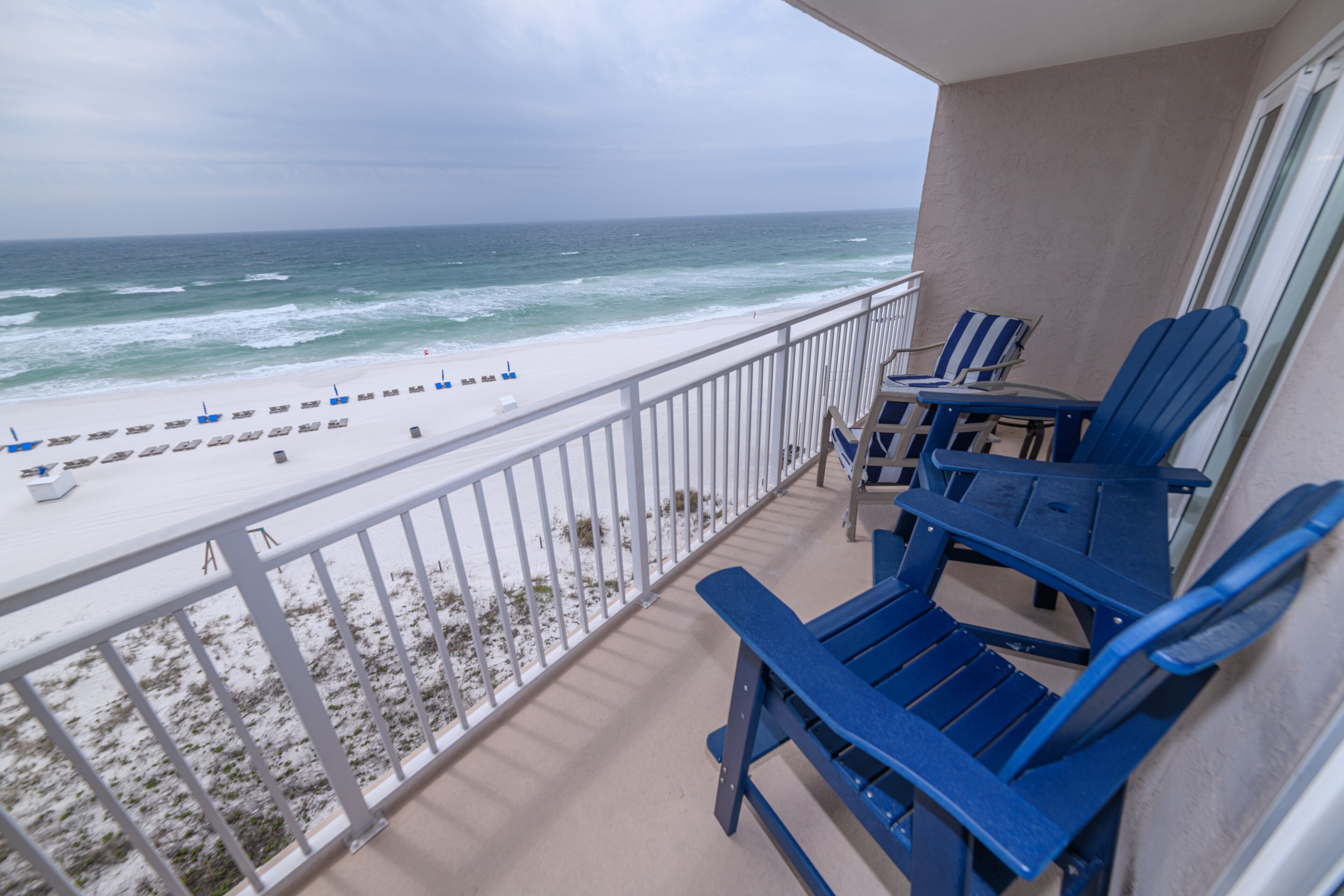 Un balcon avec des chaises bleues et une table donnant sur une plage de sable blanc, où l’on peut admirer les vagues de l’océan.