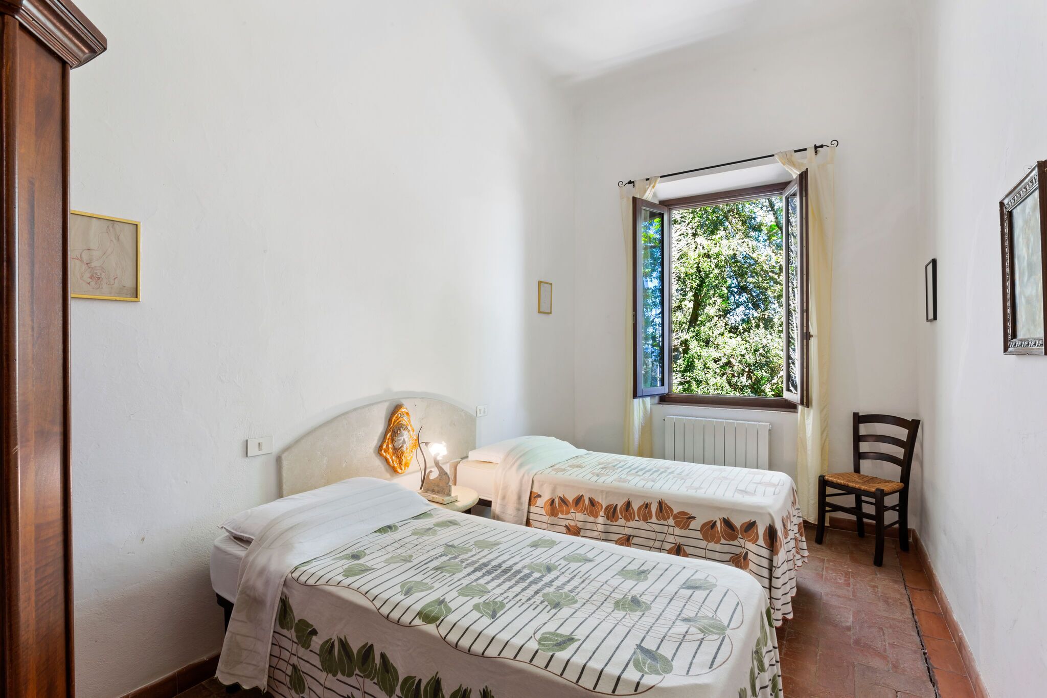 Une chambre avec deux lits, une fenêtre donnant sur les arbres, une chaise et un lampadaire fixé au mur.