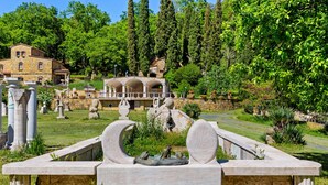 A historical site with ancient ruins, including columns and statues, surrounded by lush greenery and traditional buildings in the background.