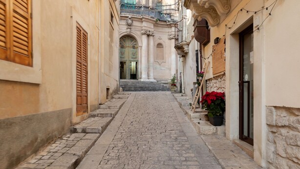 A narrow cobblestone street with buildings on either side, a set of stairs leading to a door, and potted plants. - Holiday Home in Scicli near Palazzo Ribera (Scicli)