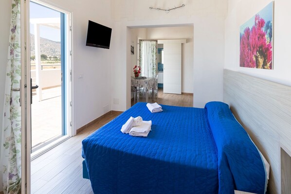 A bedroom with a blue bedspread, a flat-screen TV mounted on the wall, and a balcony door.