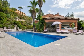 Una piscina con sillas de descanso y sombrillas, rodeada de follaje y un edificio con tejado de baldosas.