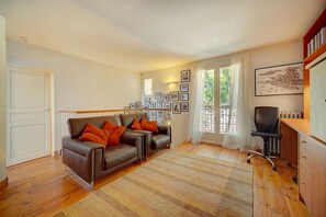 Un salon comprenant un canapé, un bureau et une chaise. Les murs sont ornés de tableaux encadrés, et il y a une fenêtre avec des rideaux.