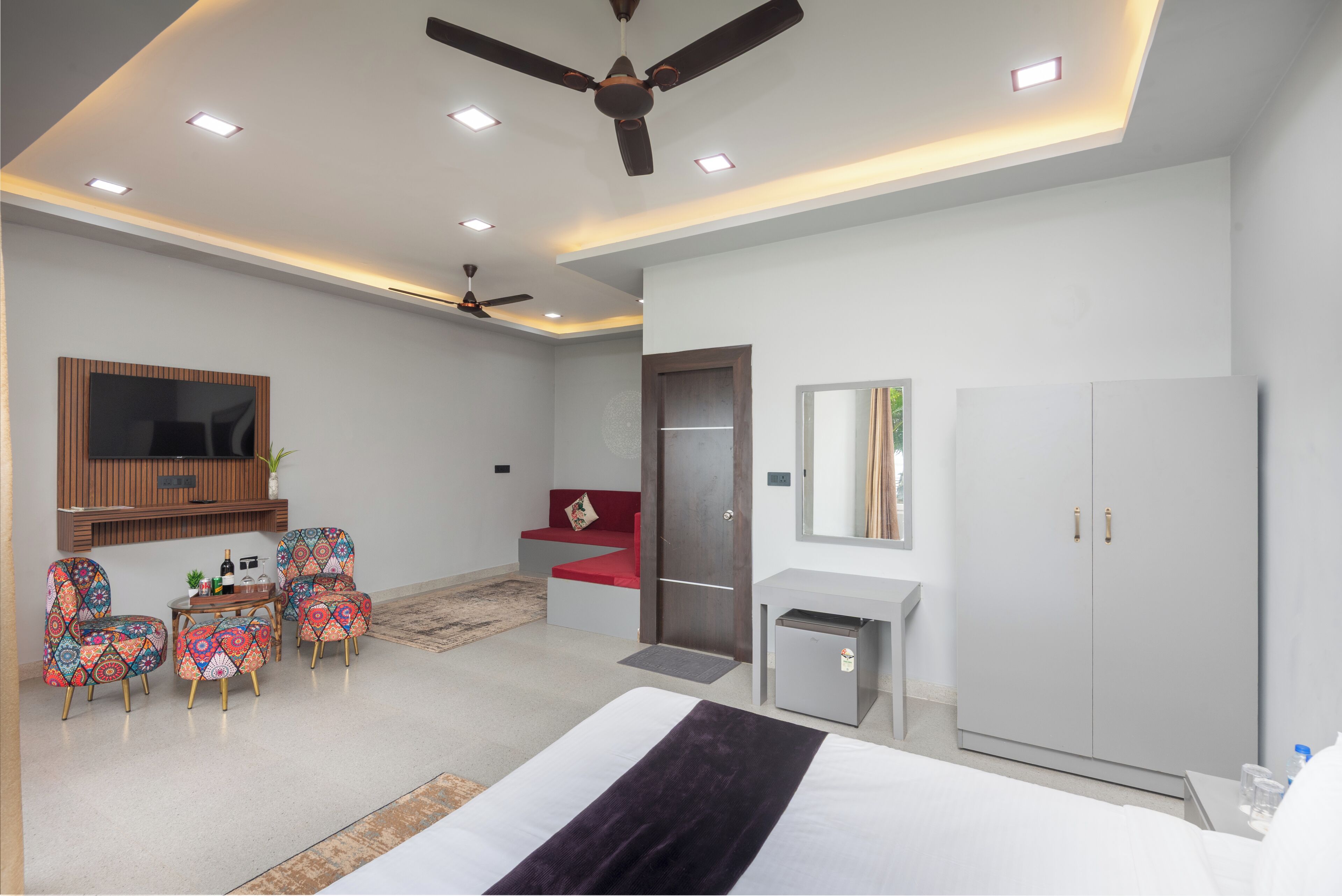 A modern hotel room with a bed, a desk, a wardrobe, a seating area with chairs, and a ceiling fan.