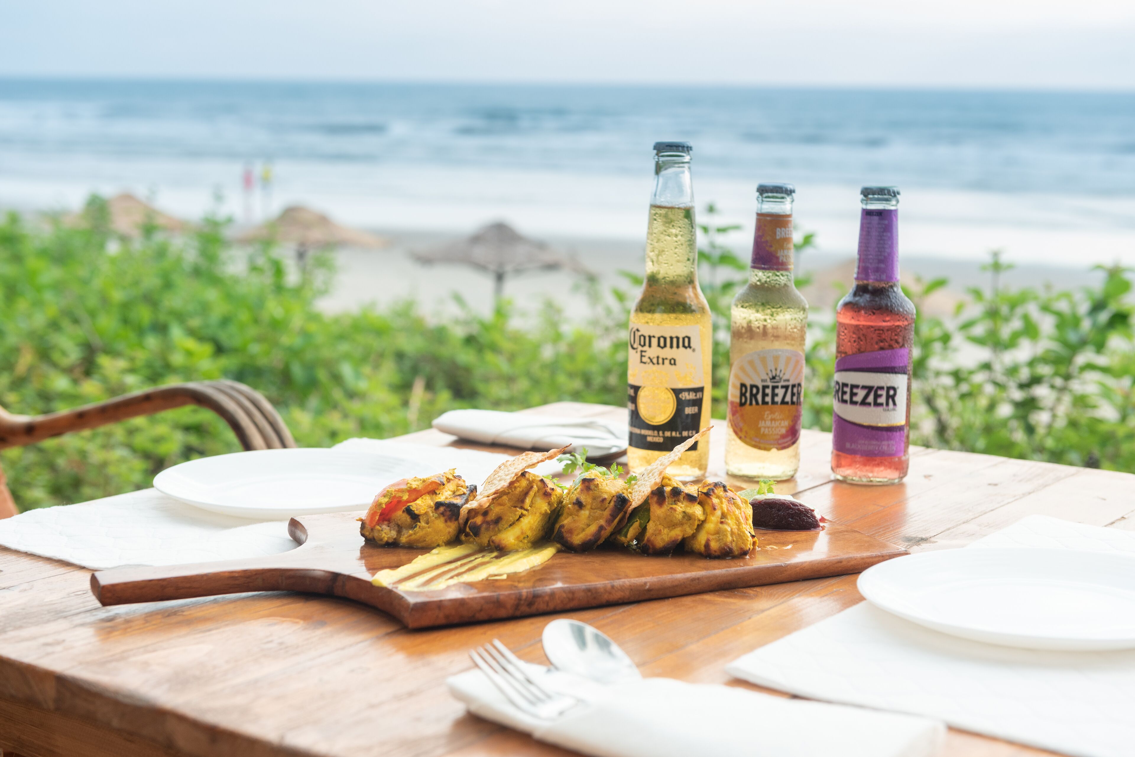 A wooden table with grilled chicken skewers, two Corona Extra beer bottles, and two BEEZER bottles, set against a beach backdrop.