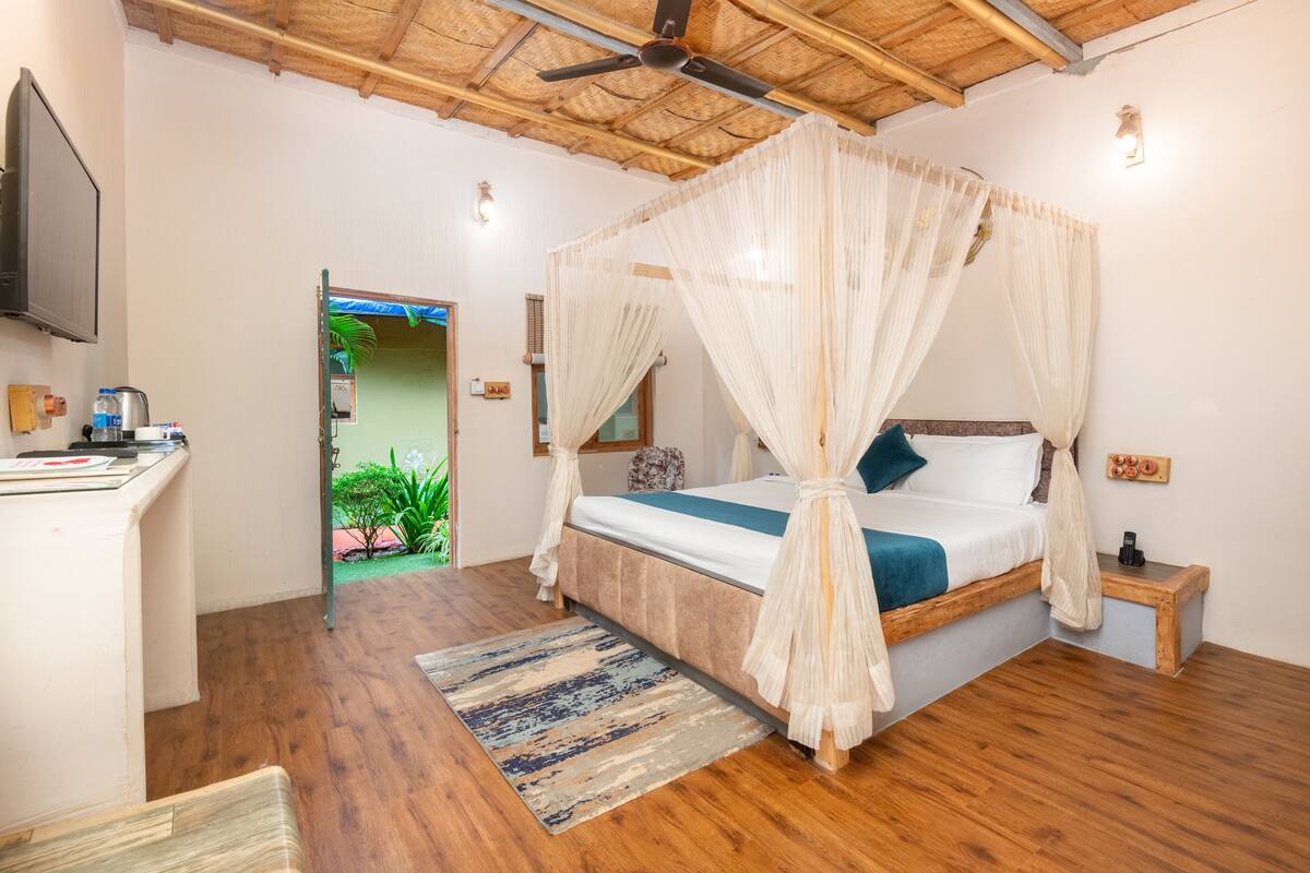 A room with a four-poster bed, wooden flooring, a ceiling fan, a TV mounted on the wall, and a small bedside table with a lamp.