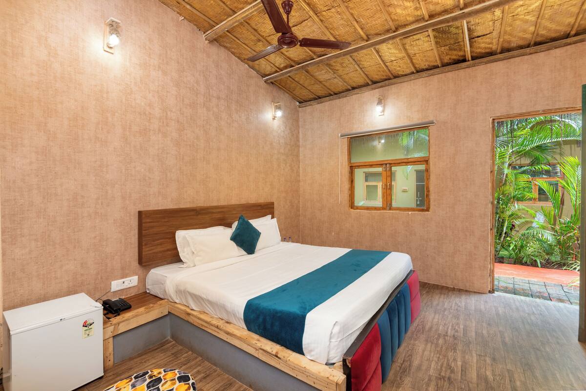 A bedroom with a bed, a small refrigerator, a ceiling fan, and a window with a view of greenery.
