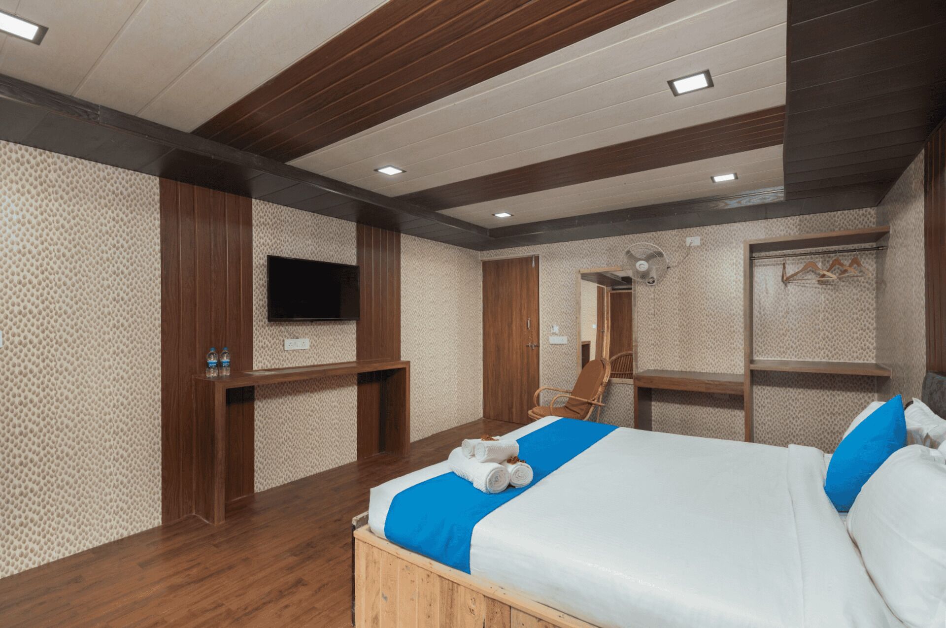 Un dormitorio con una cama de madera, un televisor montado, un espejo y un ventilador.