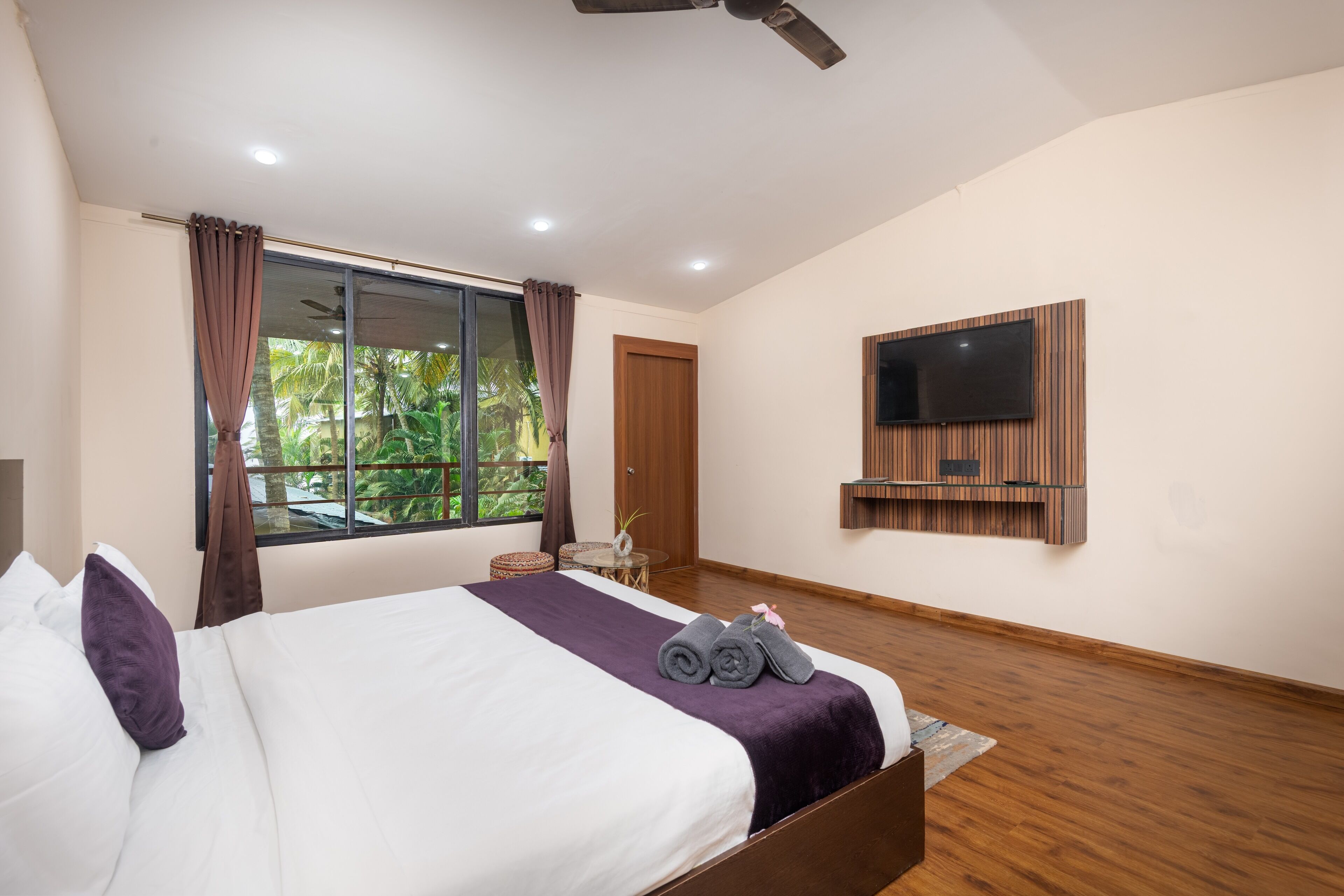 A modern hotel room with a large bed, a flat-screen TV mounted on the wall, and a view of greenery outside.