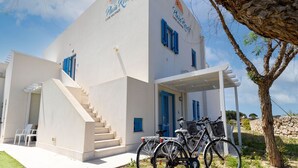Un edificio de fachada blanca, contraventanas azules y una escalera que conduce a la entrada. Hay bicicletas estacionadas afuera y un árbol cercano.