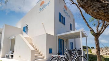 Un bâtiment à la façade blanche, aux volets bleus, avec un escalier menant à l’entrée. On peut voir des vélos garés à l’extérieur et un arbre à proximité.