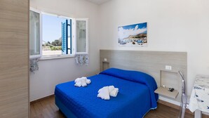 Un dormitorio con una cama grande, una ventana con contraventanas azules, un cuadro en la pared y una mesita de noche pequeña con un teléfono y una lámpara.