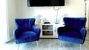 Une pièce comprenant deux fauteuils bleus, un téléviseur à écran plat fixé au mur et une console blanche agrémentée d’objets décoratifs.