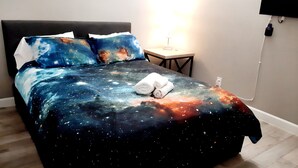 Un lit avec une couvre-lit à motifs galactiques, une lampe de chevet et un téléviseur à écran plat fixé au mur.