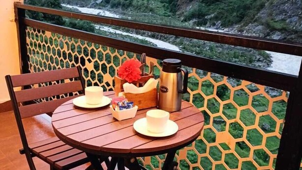Et træbord med en hvid kop, en rød blomst og en stål kaffekande på en balkon med udsigt over et bjergrigt landskab.