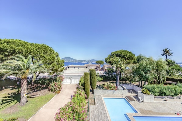A pool area with a view of the sea and mountains, surrounded by greenery and palm trees.