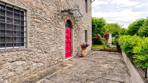 Un edificio de piedra con puerta roja, una ventana con rejas y una planta en maceta al frente.