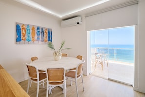 Un comedor con una mesa redonda, sillas blancas y una planta en maceta. Hay un cuadro en la pared y se ve el océano a través de una puerta de cristal.