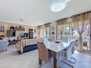 A modern living room with a dining area, a flat-screen TV, and a balcony with curtains.