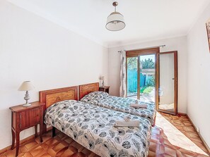 A bedroom with two beds, a nightstand, a lamp, and a sliding glass door leading to a balcony.