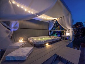 Een zone met een bubbelbad, een luifel, een tafel met lamp en een matras op het terras.