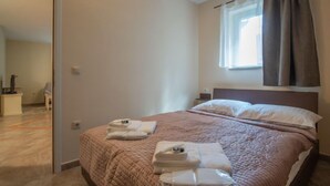 Ein ordentlich bezogenes Bett mit einer braunen Steude, ein Nachttisch mit Lampe und ein Fenster mit Vorhängen.