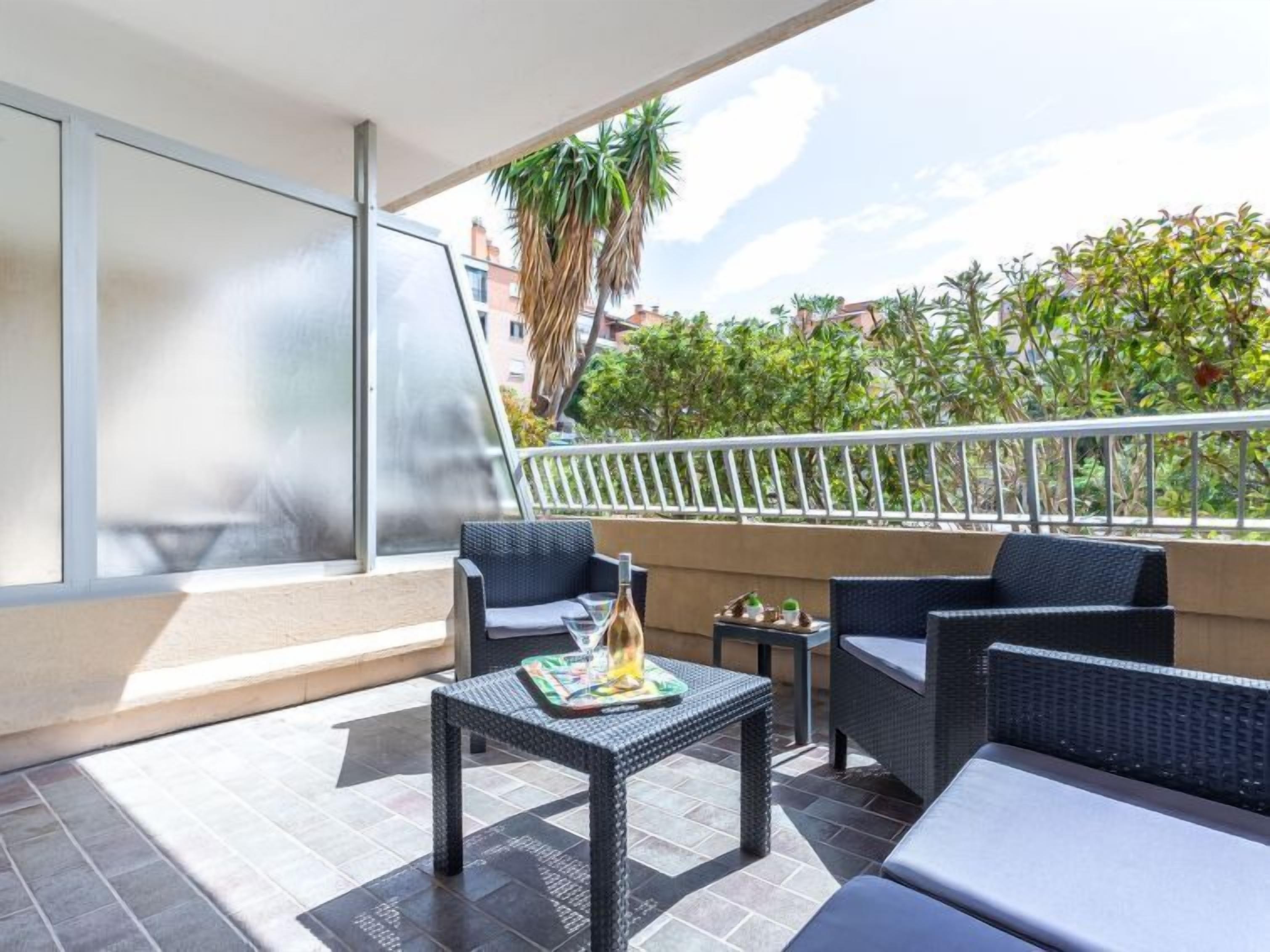 A balcony with wicker furniture, a small table with a bottle, and a view of greenery and buildings.