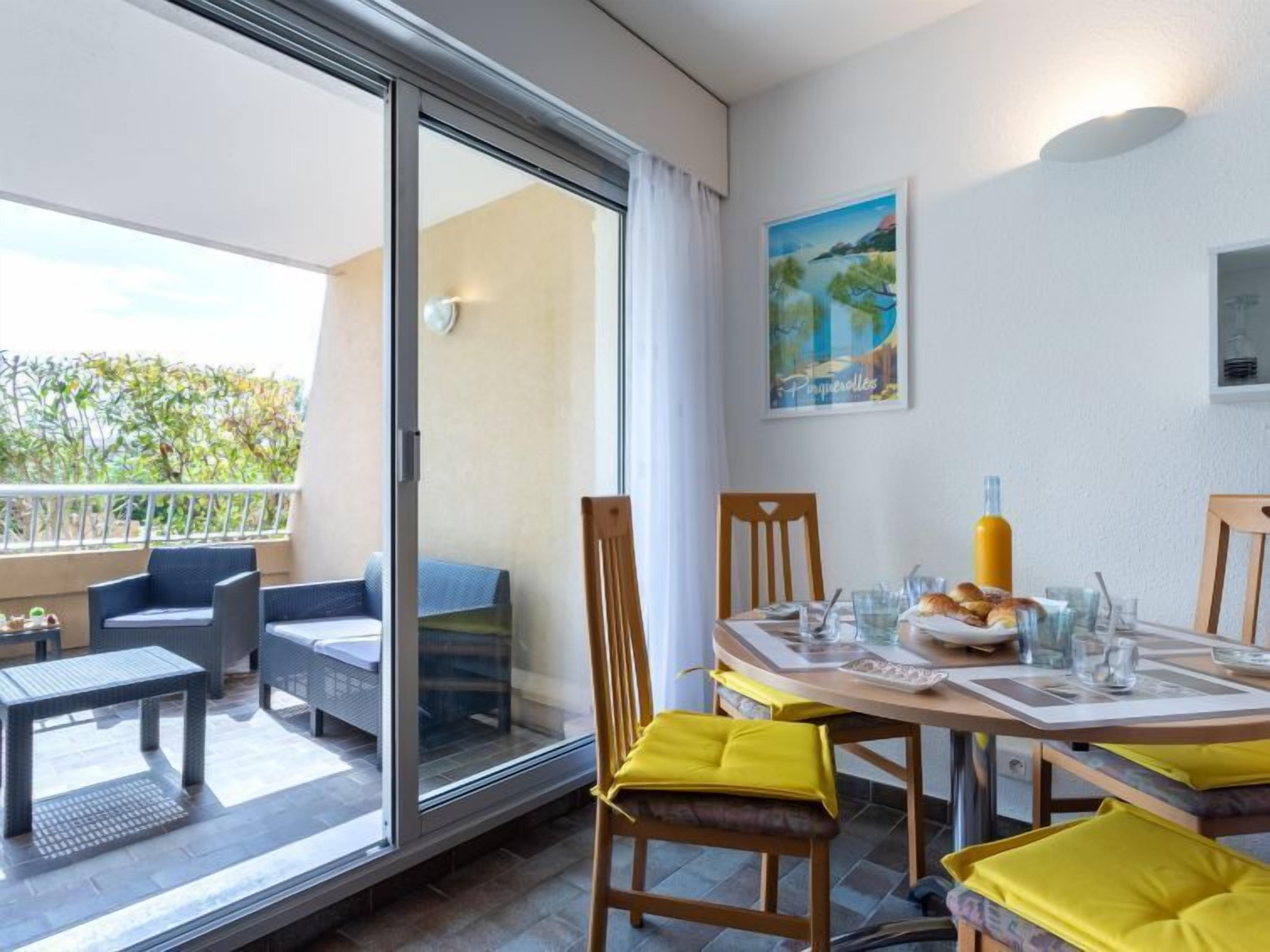 Een balkon met een glazen deur, een eettafel met gele kussens, en een schilderij aan de muur.