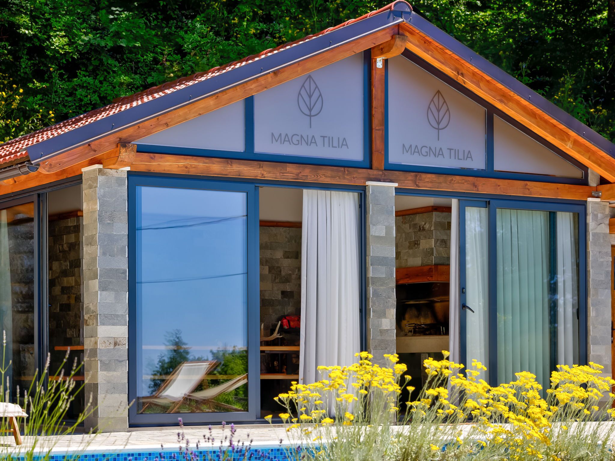 A modern building with the name "MAGNA TILIA" on the facade. It features large glass windows and doors, a stone exterior, and a pool area with yellow flowers in the foreground.