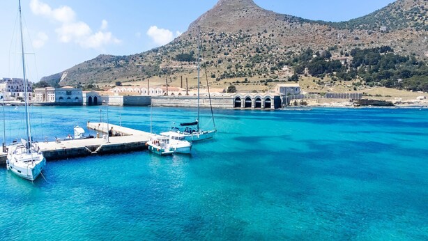 Un puerto costero con barcos amarrados, una estructura histórica en una colina y edificios con arcos.