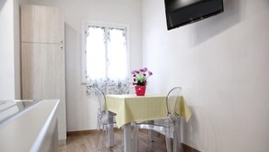 A small dining area with a table and chairs, a window with floral curtains, and a TV mounted on the wall. - Beach Home in Favignana with Pool (Favignana TP)