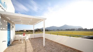 A covered outdoor area with a wooden floor, a white table with chairs, and a view of a field and mountains. - Beach Home in Favignana with Pool (Favignana TP)