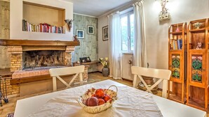 A cozy living room with a fireplace, bookshelves, a dining table with a fruit basket, and a wooden cabinet.