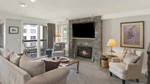 A living room with a stone fireplace, a flat-screen TV, a sofa, a chair, and a dining table. - Ski-in/Out, Walk to Base Area - Torian Plum - Pool& Hot Tubs (Steamboat Springs)
