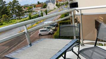 Un balcon avec une table et des chaises, donnant sur une rue bordée de voitures garées et sur laquelle on aperçoit des bâtiments au loin.
