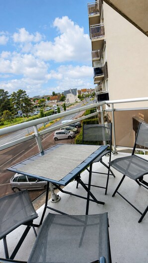 A balcony with a table and chairs, overlooking a street with parked cars and buildings in the background.