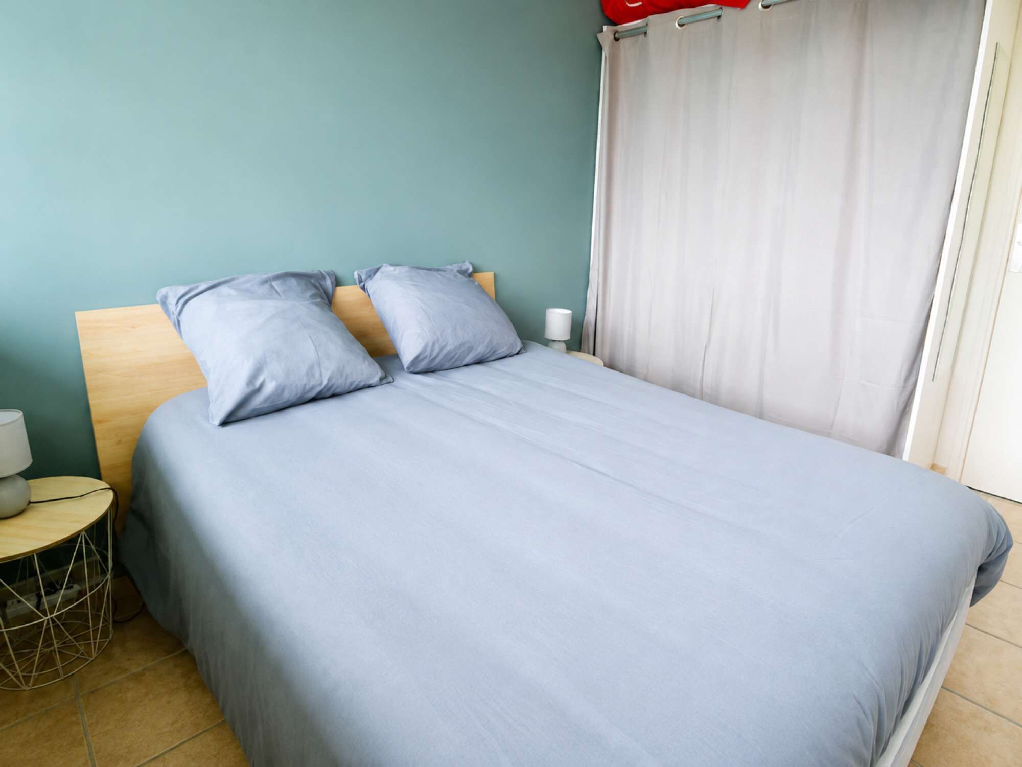 Un lit bien fait, avec une couverture et des oreillers gris, une tête de lit en bois, une table de chevet avec une lampe et un rideau à la fenêtre.