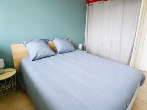 A neatly made bed with a grey bedspread and pillows, a wooden headboard, a bedside table with a lamp, and a curtain on the window.