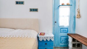 A bedroom with a bed, a blue dresser with drawers, a small table with a book and glass, and a blue door with a window. - Holiday Home in Aegina Island (Aegina Island)