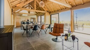 Un comedor amplio con sillas modernas y un piano, ventanas grandes que ofrecen vistas al exterior, y una estructura de techo de madera.