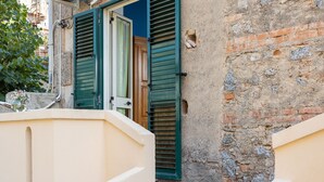 Une ruelle étroite avec un mur de couleur beige et un mur de briques, sur lequel se trouve une porte en bois et des volets verts.