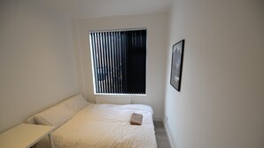 Un dormitorio pequeño y blanco con una cama individual, una ventana con persianas verticales, una mesita de noche y un cuadro enmarcado en la pared.